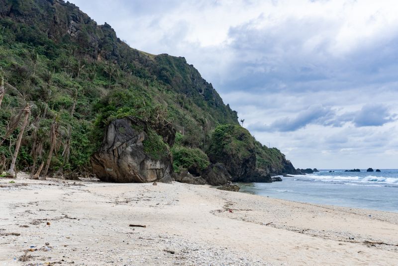 Boulders at Maydangeb White Beach. The boulders which seem to blend with the cliffs in the background are larger than the palm trees. They might be chipped off from the same cliffs through erosion rather than directly expelled via an eruption from Iraya.