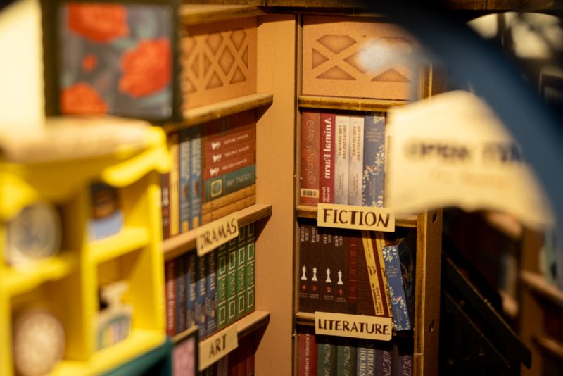 Close-up on the bookshelf behind. All labels (e.g. "FICTION") are stickers. The faux books are printed on the fiberboard directly.